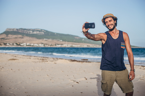 Handsome, young man with hat standing on the beach and taking selfie with mobile phone.