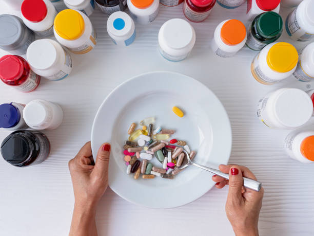 多くの食品サプリメントのペットボトルに囲まれた白いテーブルの上に座っている食品サプリメントカプセルと錠剤を持つボウルは、女性の手はそれらを食べるために錠剤の完全なスプーン� - nutritional supplement pill eating food ストックフォトと画像