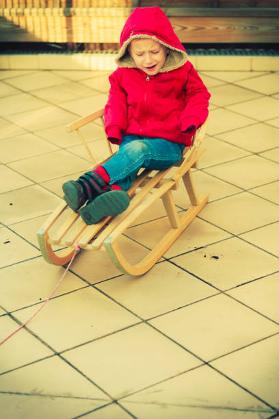 Childhood, kids imagination concept. Dissatisfied little young boy playing outside on sled but there is no snow around.