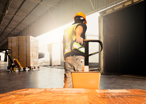 Rear view of a dock worker walking with packages on pallet jack at distribution warehouse. Young male dispatcher pulling pallet jack with boxes.