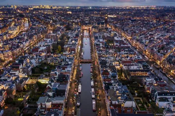 Aerial views of the Dutch capital, Amsterdam at night. Tall houses on the banks of canals with tourist boats exploring the hundreds of rivers crossing through the city.