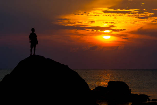 istock SILHOUETTE BOY STANDS ON HIGH ROCK LOOK AT SUNSET OVER SEA 1195400967