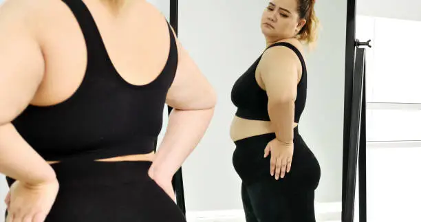Photo of Chubby woman standing and looking at her stomach in a mirror.