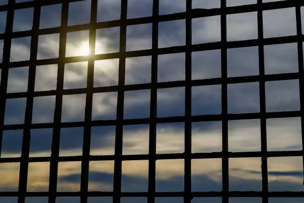 Photo of Look through the bars at the sky. Pale sunlight breaking through the dark clouds. To be imprisoned or behind the bars. To lose freedom concept. Hopelessness and loneliness.