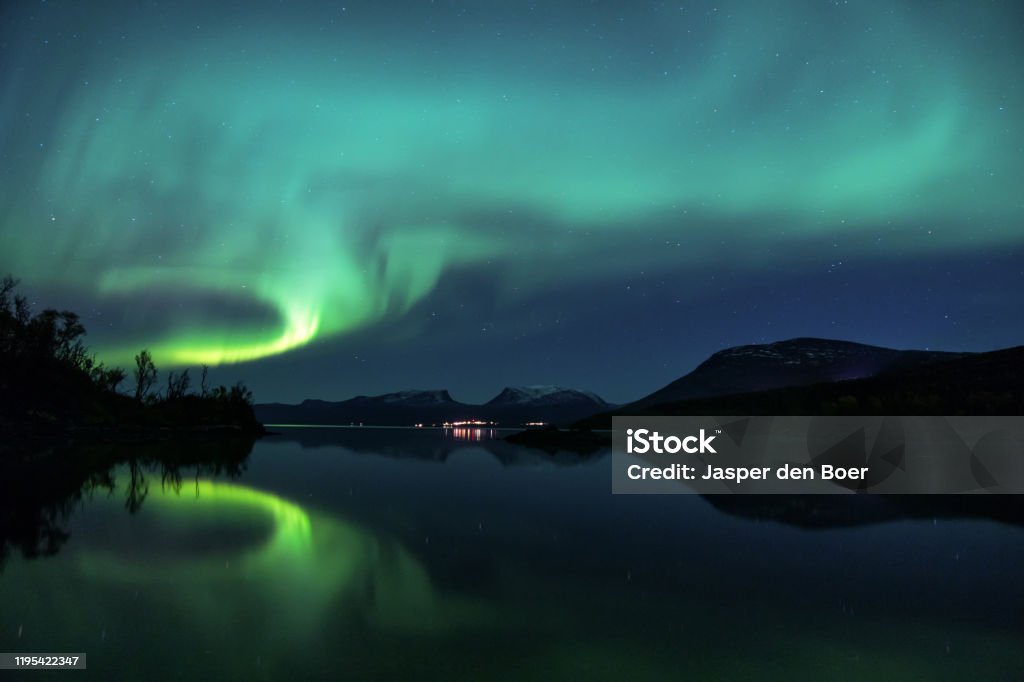 Northern Lights reflecting in the water in Abisko, Sweden A beautiful Aurora Borealis display, reflecting in Lake Torneträsk, over Abisko in Swedish Lapland. Above the city lights, you can see the iconic 'Lapporten', a characteristic valley symbolising the gate to Lapland. Abisko National Park Stock Photo