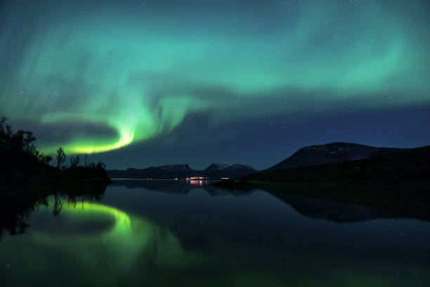 Northern Lights reflecting in the water in Abisko, Sweden A beautiful Aurora Borealis display, reflecting in Lake Torneträsk, over Abisko in Swedish Lapland. Above the city lights, you can see the iconic 'Lapporten', a characteristic valley symbolising the gate to Lapland. norrbotten province stock pictures, royalty-free photos & images