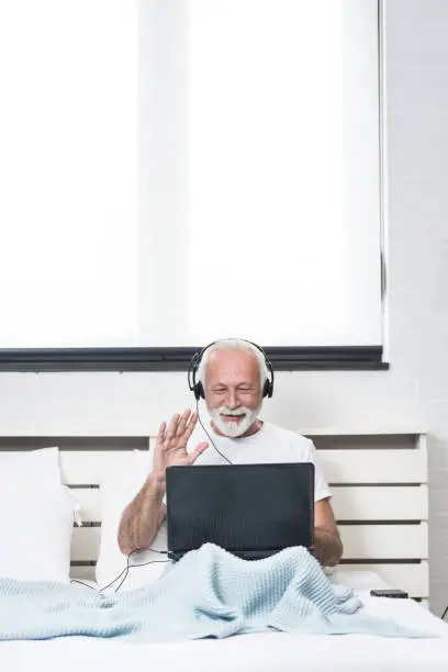 Senior grey haired man doing video call from his laptop while sitting on his bed with ear headset. Copy space on white background