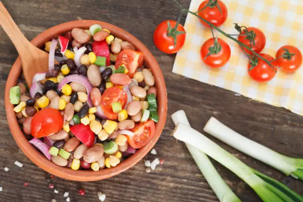 Vegetarian salad with red and black beans, chickpeas and corn in clay bowl on rustic wooden table. Healthy country food with mixed vegetables.