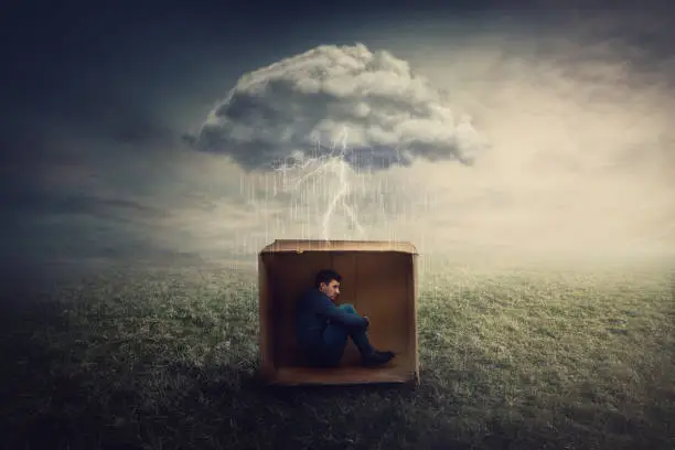 Photo of Surreal concept with a scared guy shelters inside a cardboard box. Introvert man caged by own fears as a thunderstorm cloud trapped him under the rain. Mysterious storm as emotional crisis symbol.