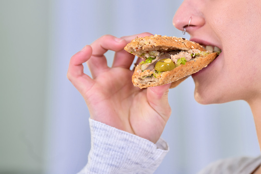 Close-up of a woman biting a delicious looking tuna sandwich with olives and anchovies on an out of focus light background.