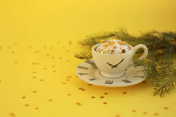 Ceramic white cup with glitter stars inside and small saucer with clock face icon, evergreen tree branch. Yellow background.