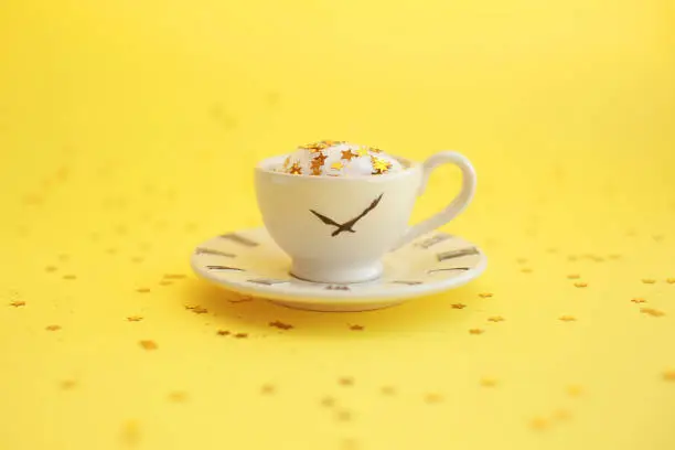 Ceramic white cup with glitter stars inside and small saucer with clock face icon. Yellow background.