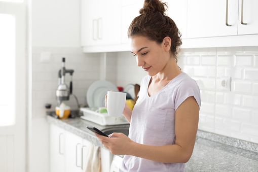 Young woman holding coffee and using mobile phone at kitchen