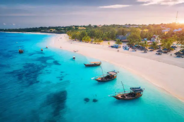 Photo of Aerial view of the fishing boats on tropical sea coast with white sandy beach at sunset. Summer holiday on Indian Ocean, Zanzibar. Landscape with boat, palm trees, transparent blue water. Top view