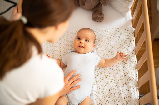 Mother leaning over side of crib and talking to her baby boy. Child looking at mum and smiling