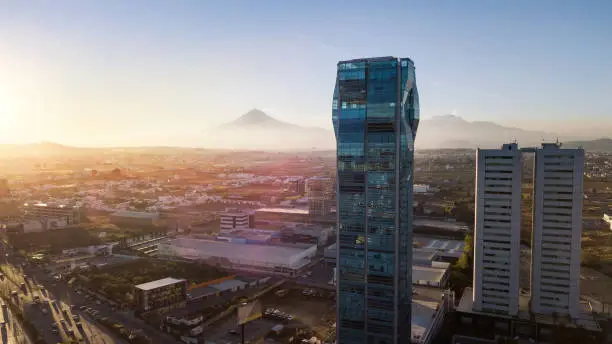 Aerial view of the rapidly urbanizing area of Puebla City, Mexico, where skyscrapers are being constructed at a hurried pace, while the Popocatépetl volcano looms in the distance.