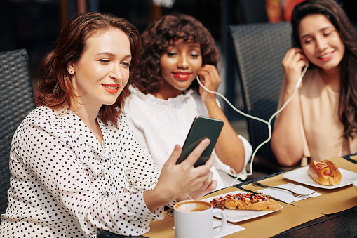 Smiling young woman turning on music track on her smartphone and giving earphones to friends