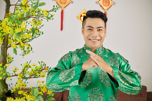 Happy smiling Vietnamese young man in traditional costume making special gesture to wish happy Lunar New Year