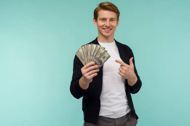 belo cara de cabelos vermelhos desportivo apontando dinheiro do dedo em um fundo azul. - holding money - fotografias e filmes do acervo