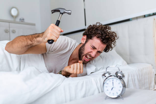 젊은 남자는 망치로 알람 시계를 깰 시계를 파괴하려고합니다. 침대에 누워있는 남자는 아침 7시에 망치로 알람 시계를 끄고 있습니다. - wake 뉴스 사진 이미지