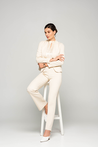 elegante mujer de negocios confiada en traje blanco sentado en el taburete, en gris photo