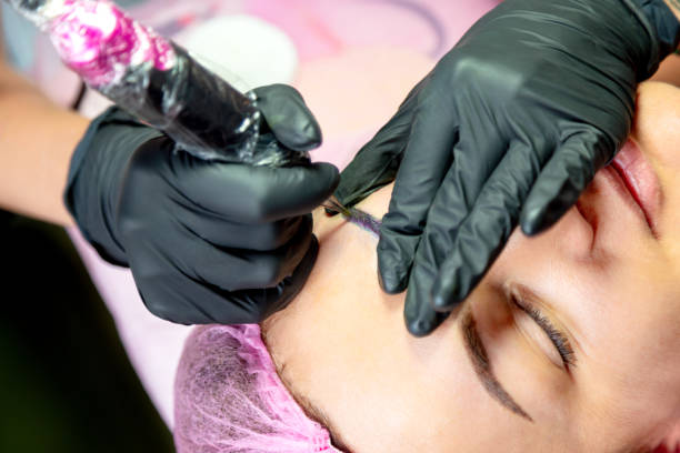 istock Cosmetologist making permanent makeup - microblading eyebrows. Woman getting facial care and tattoo at beauty salon. Tattooing machine and hair is in protective wear. 1195543583