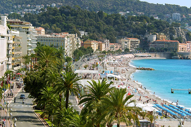 Nice, France - July 20, 2011. The seaside promenade with the „Walkway of the English“ (Promenade des Anglais) in summer time. The beach is overcrowded with people from all parts of the world. The Maritime Alps in the background and palm trees in front. Bird's eye view from the cathedral hill (Colline du Chateau).