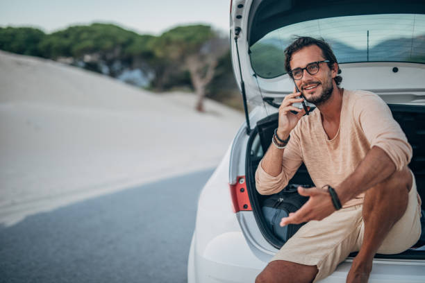 istock Handsome man sitting on car trunk and talking on mobile 1195554796