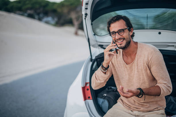 istock Handsome man sitting on car trunk and talking on phone 1195554815