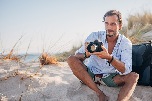 One man, handsome man sitting on the beach, taking pictures with camera.