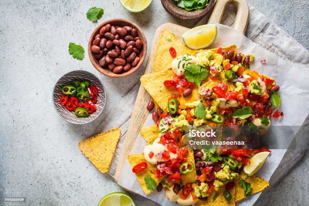 Nachos chips with cheese sauce, guacamole, salsa and vegetables on the board, top view. Party food concept. Nachos chips with cheese sauce, guacamole, salsa and vegetables on the board. Party food concept. Mexican food concept. Nacho Chip Stock Photo