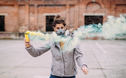 Street artist wearing mask. Hipster girl playing with yellow smoke bomb.