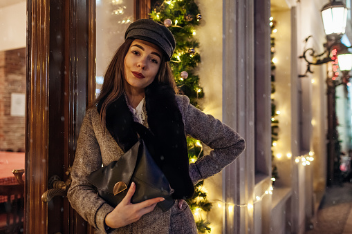 Christmas, New Year winter fashion. Woman walking on city street by decorated showcases. Stylish girl holding purse under falling snow