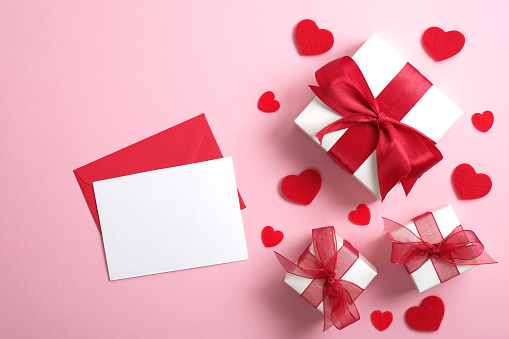 Love Letter with blank paper note and white gift boxes with red ribbon bow on pastel pink background decorated red hearts. Flat lay, top view. Saint Valentine's Day surprise, love concept.