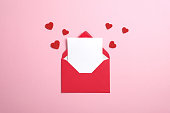 内側に白いノートモックアップが入った赤い紙の封筒と、ピンクの背景にバレンタインハート。フラットレイ、トップビュー。バレンタインデーのコンセプトのためのロマンチックなラブレ�