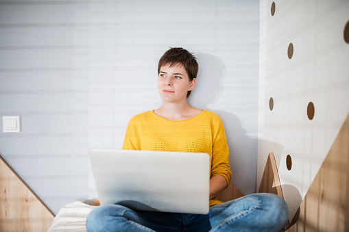 Front view of young woman with laptop sitting on bed in bedroom indoors at home.