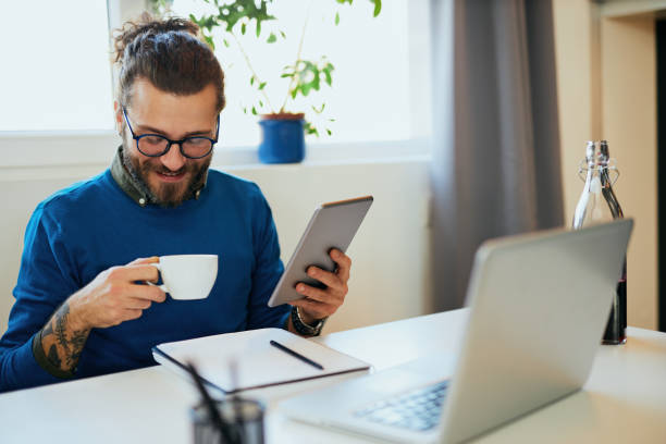 istock Young pensive attractive caucasian bearded freelancer sitting in his home office, using tablet for reading news and holding cup of coffee. 1195618271