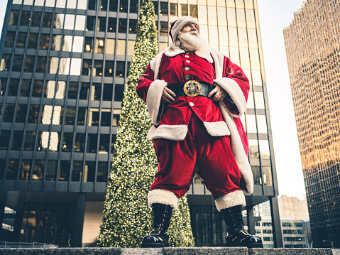 Santa Clause posing in front of large Christmas tree in big North American city. Long white beard, Red suit, boots and all. Ready for Christmas.