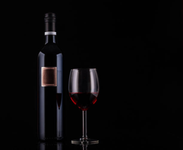 Closed red wine bottle with empty label and glass of wine on black background with reflections. Concept sales, discount price.