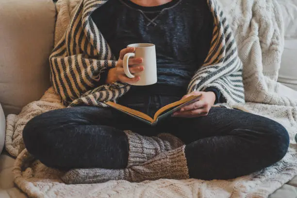 A woman drinking coffee on a sofa while reading a book.