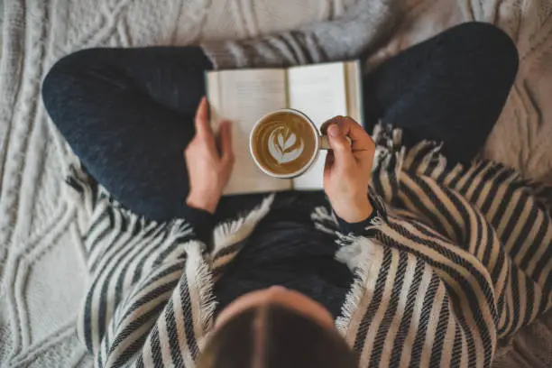 A woman drinking coffee on a sofa while reading a book.