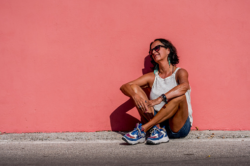 Attractive older woman with suntan wearing sunglasses leaning against a bright light pink monochrome wall. Sunny sunshine day one person older mature female.
