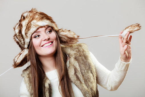 Winter fashion. Happy young woman wearing fashionable wintertime clothes fur cap studio shot on gray background