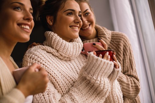 Low angle view of three happy female friends in woolen clothes holding cups and looking something through the window.