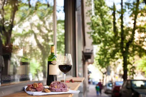Photo of slices of baguette bread, brie cheese and saucisson (a cured meat from France) for appetizers with a Glass and bottle of French red wine, blurred in the background, on the table of a terrace of Paris.