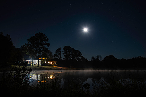 Beautiful residential house in the country with interior lights on, reflected in the nearby lake on a moonlit night.