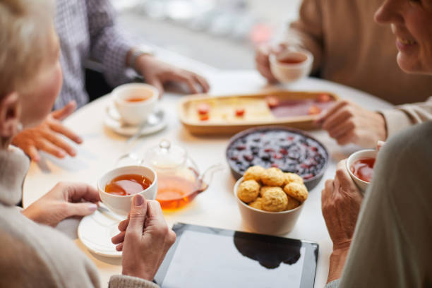 vue au-dessus d'épaule des personnes âgées s'asseyant à la table avec le comprimé et buvant le thé avec des sucreries tout en parlant les uns aux autres - tea party party tea little girls photos et images de collection