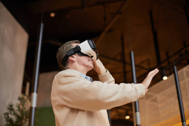 istock Below view of concentrated senior man in virtual reality goggles gesturing hand while playing video games 1195697905