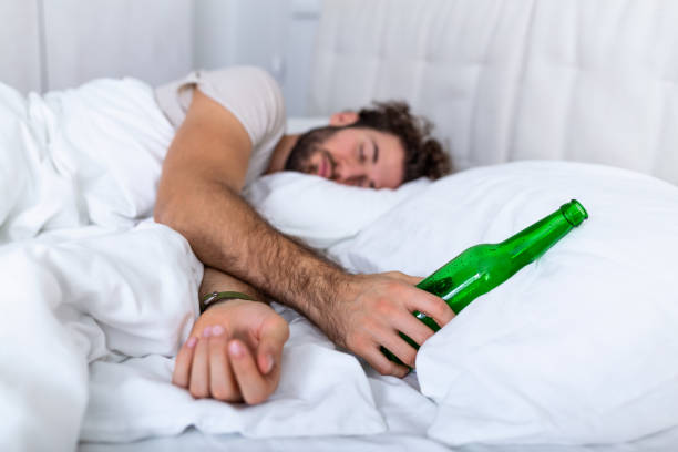drunk man in the bed and sad place and an alcohol bottle in his hand. young man lying in bed deadly drunken holding near-empty bottle of booze. - eastern european caucasian one person alcoholism imagens e fotografias de stock