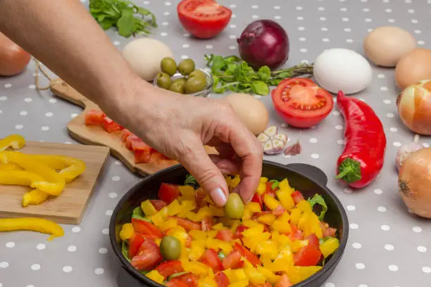 Photo of Sliced vegetables in pan. Hands put  olive in pan. Olives, eggs,